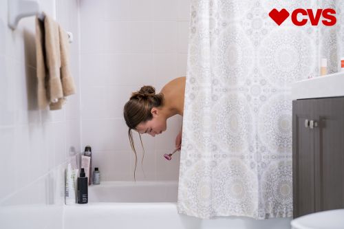 Woman in a white tiled bathroom bending at the waist, holding a pink facial razor, next to a white and grey patterned shower curtain.