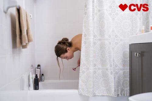 Woman in a white tiled bathroom bending at the waist, holding a pink facial razor, next to a white and grey patterned shower curtain.