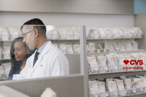Two pharmacy employees, an adult male and an adult female, stand inside a CVS pharmacy behind a glass barrier, with shelves of product visible.