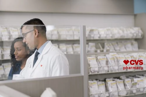 Two pharmacy employees, an adult male and an adult female, stand inside a CVS pharmacy behind a glass barrier, with shelves of product visible.