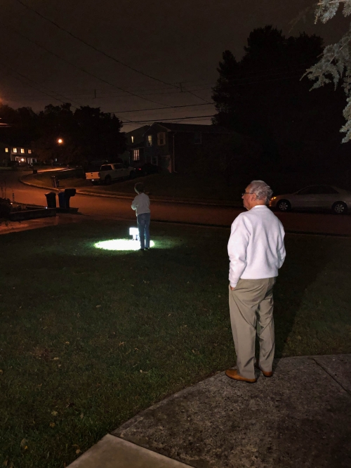 Two people outside at night with spotlight on the grass in a suburban neighborhood with houses and parked cars visible in the background.