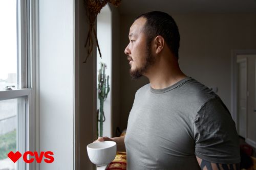 Adult male with a slight beard in profile holding a white coffee cup looking out of a bright, daylight window inside a home.