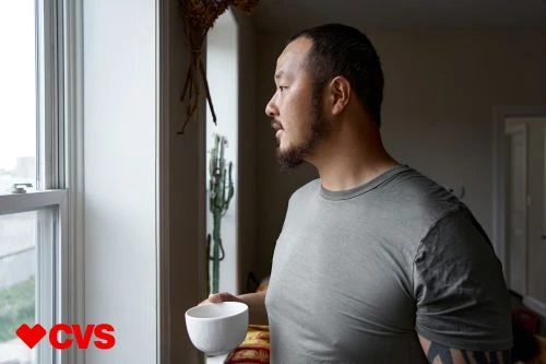 Adult male with a slight beard in profile holding a white coffee cup looking out of a bright, daylight window inside a home.
