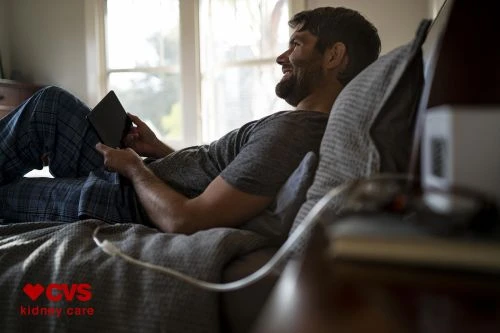 Man with beard lying in bed wearing plaid pajama pants and a grey t-shirt while holding a digital tablet