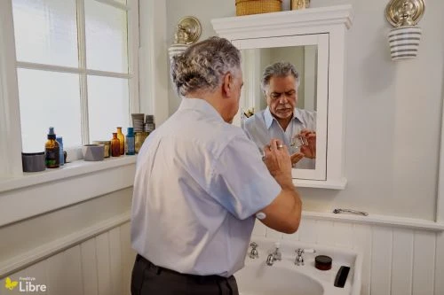 Mature adult male in a bathroom, looking into a mirror and adjusting eyeglasses, in a home interior with natural lighting and a white sink.