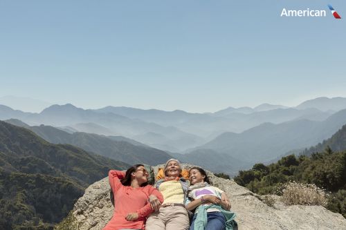 Three people, likely family members, lounge on a rock outcropping, overlooking a scenic mountain range with clear sky in the background.