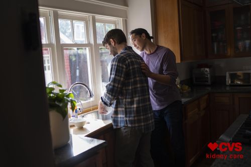 Two men in a kitchen, one washing dishes at the sink near a window while the other places a hand on his shoulder.