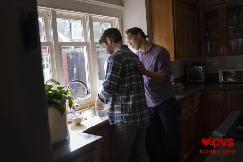Two men in a kitchen, one washing dishes at the sink near a window while the other places a hand on his shoulder.