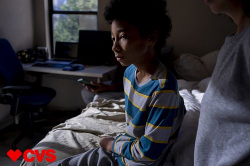 A child sits on a bed in a bedroom with an adult in the foreground. The child is wearing a striped shirt and grey pants.