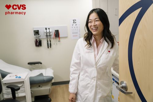 Smiling Asian woman in a white doctor's coat stands in a medical exam room with an exam table, medical equipment, and eye chart visible.