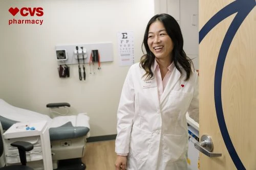 Smiling Asian woman in a white doctor's coat stands in a medical exam room with an exam table, medical equipment, and eye chart visible.
