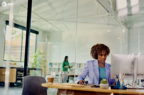 Professional African-American woman working at her computer in a modern office, wearing a blue blazer. Glass wall reflects second person.