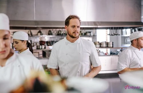Professional kitchen scene features three chefs in white uniforms and hats, with focus on one male chef, set against stainless steel backdrop.