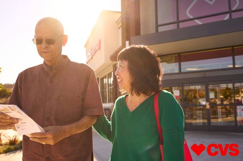 Elderly Asian couple outside CVS Pharmacy. Man holds paper, both wear casual attire in bright sunlight near store entrance.