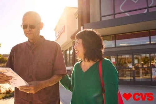 Elderly Asian couple outside CVS Pharmacy. Man holds paper, both wear casual attire in bright sunlight near store entrance.