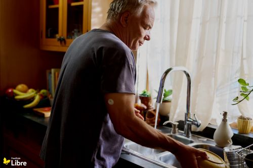 Mature man washing dishes in a bright kitchen, wearing a grey shirt, with house plants and fruit visible in the background.