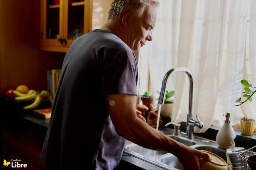 Mature man washing dishes in a bright kitchen, wearing a grey shirt, with house plants and fruit visible in the background.
