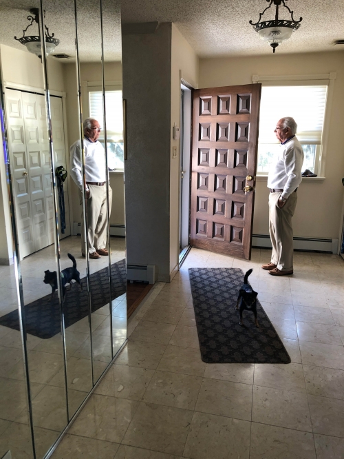 Elderly man standing near an open door with a black cat on a tiled floor, reflection in mirrored closet doors.