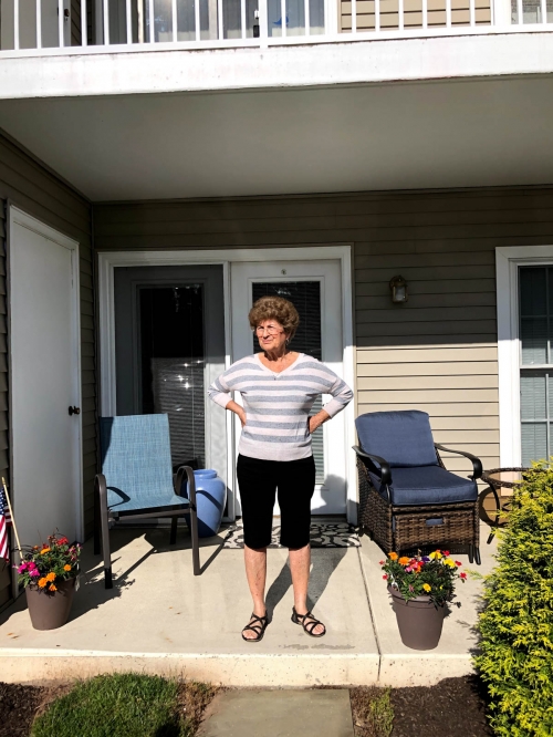Outdoor portrait of an elderly woman standing on a porch in front of a building with flowerpots and chairs.