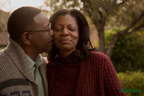 African American couple shares an intimate moment outdoors. The man kisses the woman's cheek as she smiles gently, framed by soft natural light.