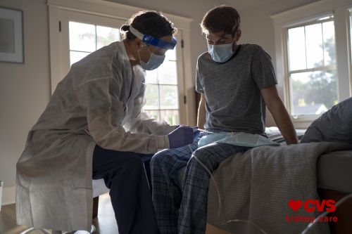 In-home healthcare visit: A healthcare worker wearing PPE administers a medical test to a patient sitting on a bed in a home interior.