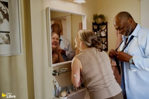 An older couple smiling in a bathroom, with a man tying his shirt and a woman looking into a mirror.