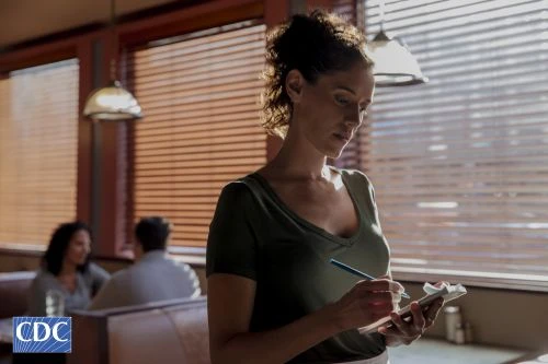 Woman waitress taking order with notepad and pen in a restaurant interior with blinds and other customers in the background.