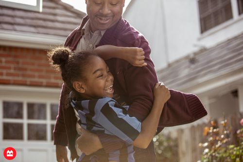 A smiling black father lifting his joyful young daughter outdoors in front of a residential house.