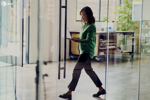 Professional woman walks through a glass door in an office while holding a laptop; natural light fills the workspace.