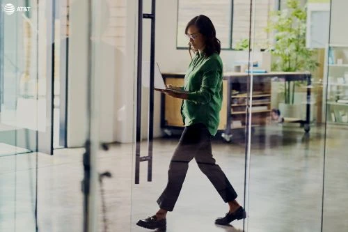 Professional woman walks through a glass door in an office while holding a laptop; natural light fills the workspace.
