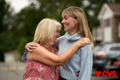 Two smiling women embracing in a driveway outdoors, with a house and cars visible in the background, and a CVS logo in the corner.