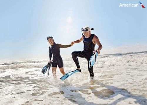Active senior couple in wetsuits walk on sandy beach toward water with scuba gear, enjoying their vacation with airline logo.
