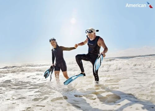 Active senior couple in wetsuits walk on sandy beach toward water with scuba gear, enjoying their vacation with airline logo.