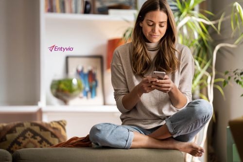 Young woman sitting cross-legged on an ottoman indoors, looking at her smartphone, with houseplants and a shelf in the background.