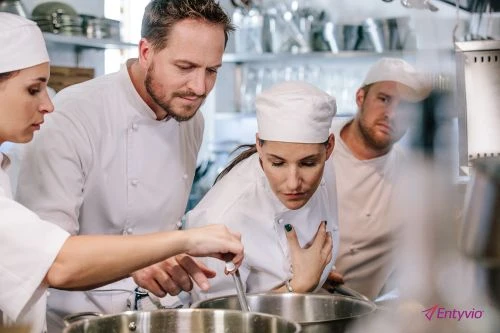 Four chefs in white uniforms and hats focus on stirring a pot on a stove in a commercial kitchen with stainless steel equipment.