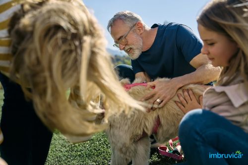 Senior man and young girl petting a dog outdoors on a sunny day, family bonding with their golden retriever, Imbruvica logo.