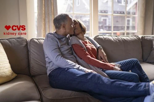 Mature couple shares a kiss while sitting on a couch indoors near a window, with the CVS Kidney Care logo visible in the image.