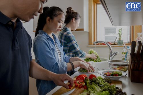 Three Asian people preparing a healthy salad in a bright kitchen near a window, showcasing fresh produce and a clean culinary environment.