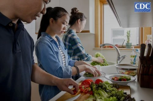 Three Asian people preparing a healthy salad in a bright kitchen near a window, showcasing fresh produce and a clean culinary environment.