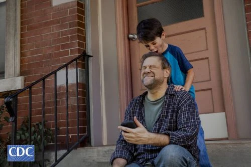 A father and son interact playfully outdoors on the front steps of a brick house. The child is playfully covering the dad's eyes.