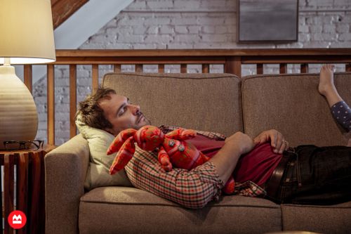 Man lying asleep on a brown sofa holding a red stuffed toy rabbit, photographed by Augustus Butera