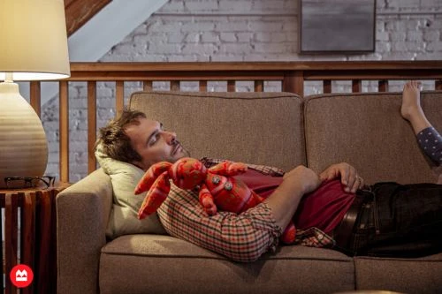 Man lying asleep on a brown sofa holding a red stuffed toy rabbit, photographed by Augustus Butera