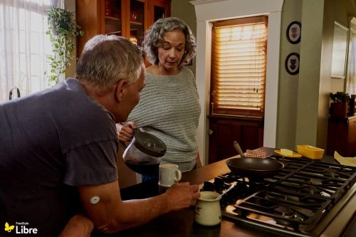 Mature couple at home in kitchen pouring coffee, preparing breakfast near the stove with natural window light indoors during daytime.
