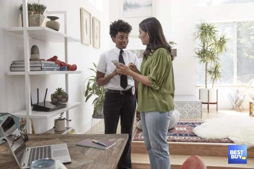 Two women in a brightly lit home interior are looking at a smartphone. One woman wears a uniform, and the other wears casual clothing.