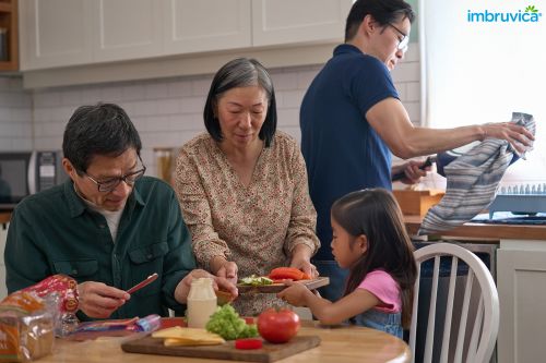 Family makes sandwiches together in a bright kitchen: child, adult male and female, gray cabinets, wooden table, Imbruvica logo.