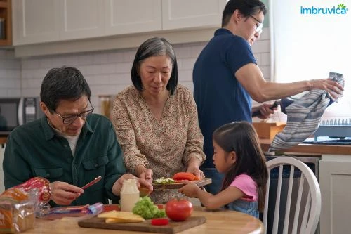 Family makes sandwiches together in a bright kitchen: child, adult male and female, gray cabinets, wooden table, Imbruvica logo.