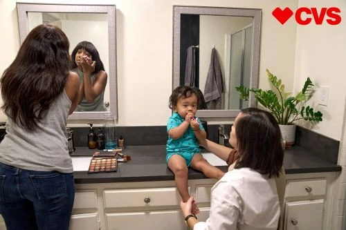 Woman in grey tank top and jeans looking in a bathroom mirror, while another woman helps a toddler seated on a bathroom counter.