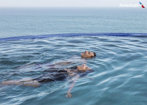Couple relaxing in infinity pool overlooking the ocean. Serene travel and lifestyle scene with an American Airlines logo in the corner.