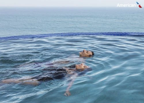 Couple relaxing in infinity pool overlooking the ocean. Serene travel and lifestyle scene with an American Airlines logo in the corner.