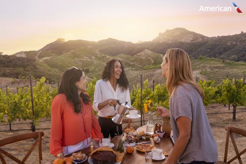 Three women enjoy outdoor dining at a vineyard, featuring fresh food, drinks, and a scenic mountain backdrop at sunset.